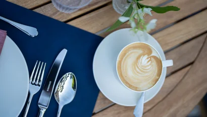 Kaffee mit Milchschaum auf einem weißen Teller neben Besteck auf blauem Tischset